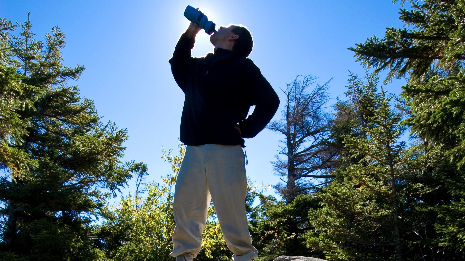 A man drinking from his water bottle while on a hike with the sun beaming behind him on a warm autumn day