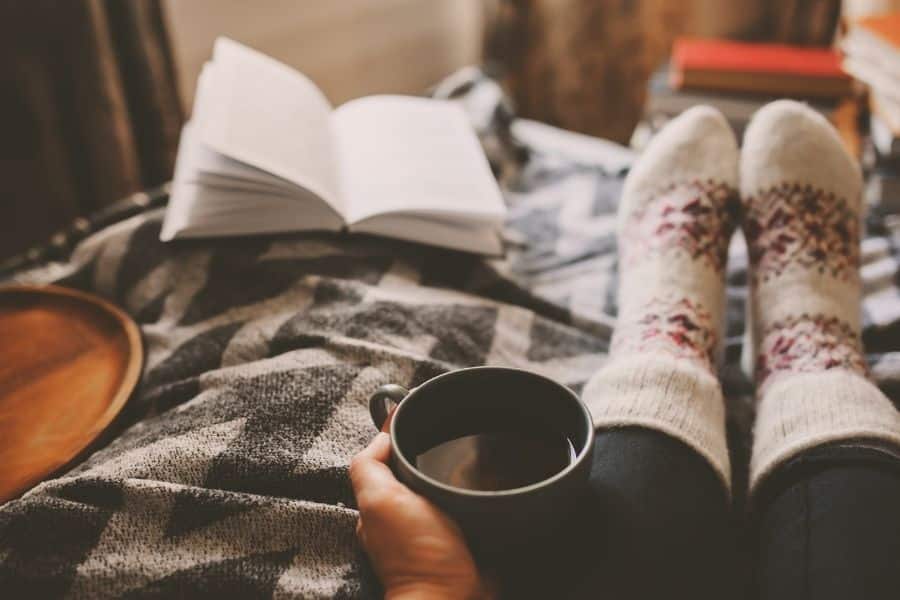 someone with cozy socks sitting on a wamr blanket by a fire with an open book and mug of hot cocoa