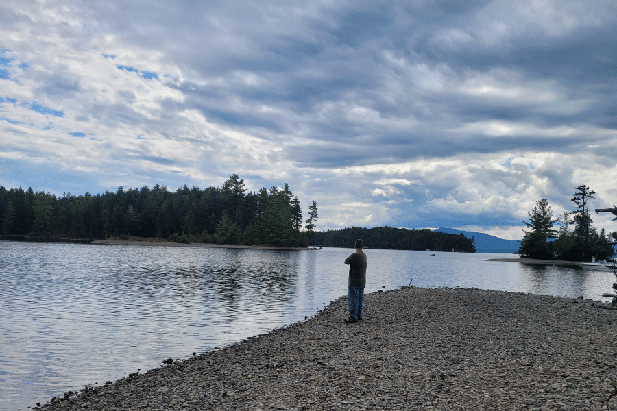 Man standing on a rocky shoreline at Lily Bay State Park, overlooking Moosehead Lake surrounded by pine-covered islands under a partly cloudy sky in Maine.
