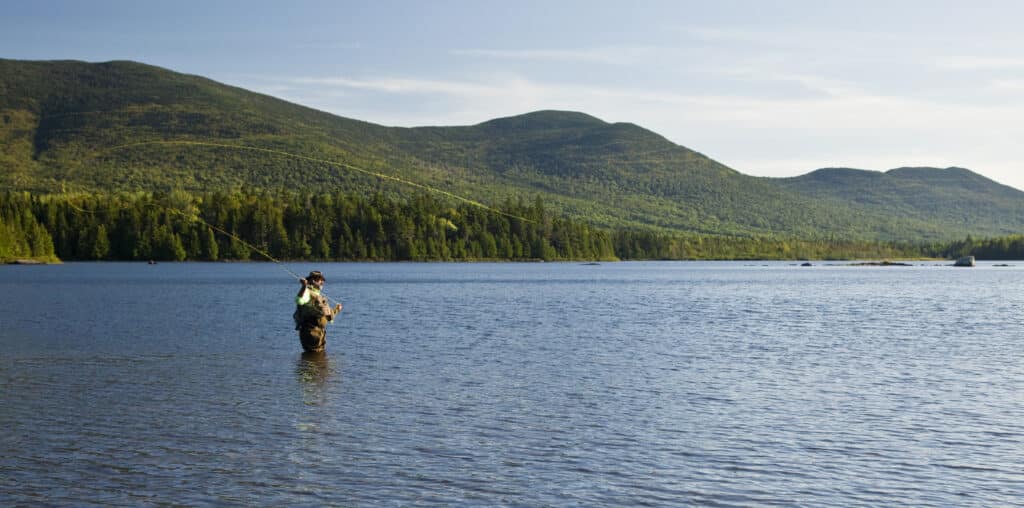 Fly fisherman casting a line in Moosehead Lake with forested mountains in the background, showcasing the scenic outdoor adventure near Lodge at Moosehead Lake in Maine.