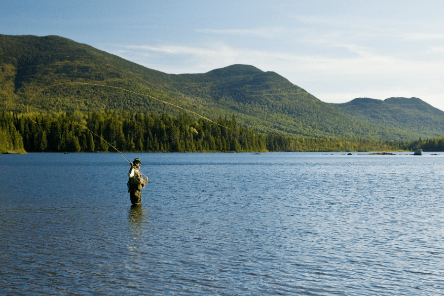 Fly fisherman casting a line in Moosehead Lake with forested mountains in the background, showcasing the scenic outdoor adventure near Lodge at Moosehead Lake in Maine.